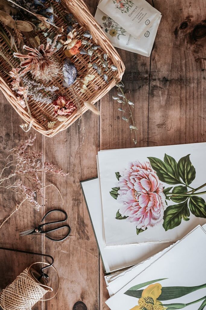 pexels photo 9350677 Top view of botanical paintings with dried flowers, scissors, and basket on a wooden surface, creating a cozy artistic vibe.
